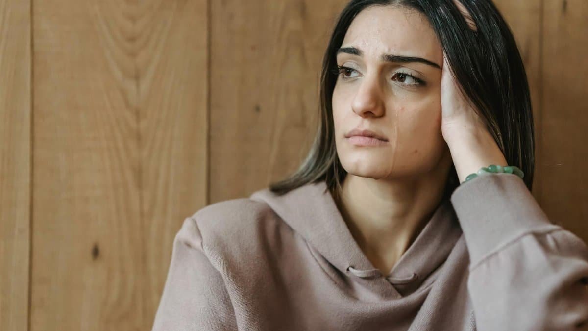 Unhappy young woman with brown hair in casual clothes leaning on hand and looking away while crying in room with wooden wall