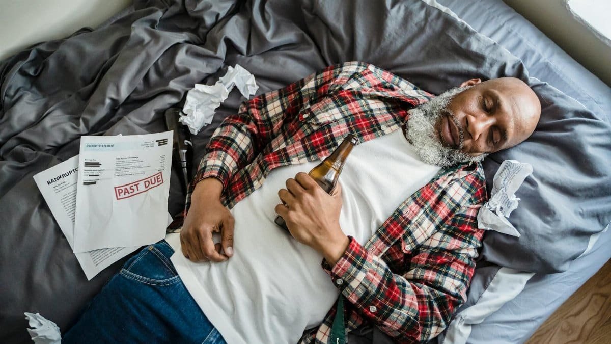 A senior man asleep on a bed, surrounded by past due bills, holding a beer bottle.