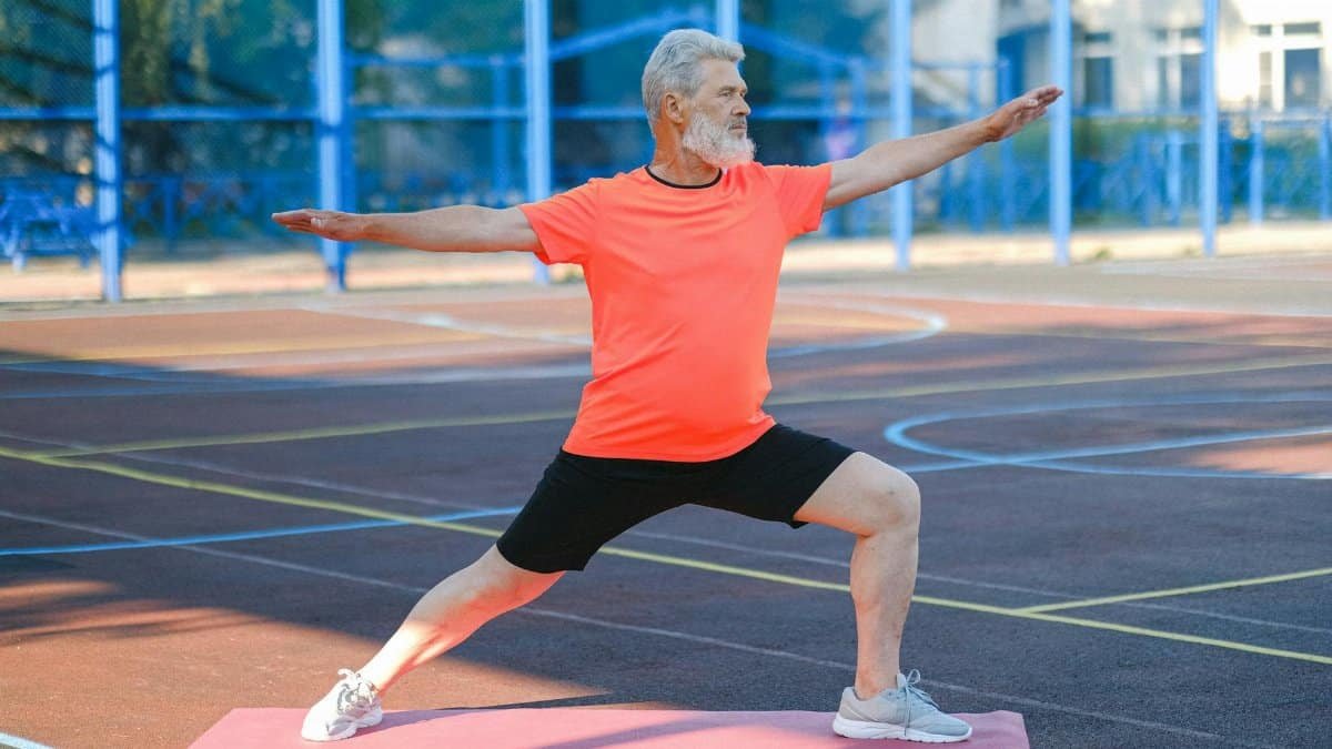 Elderly man performing warrior II yoga pose on an outdoor court.
