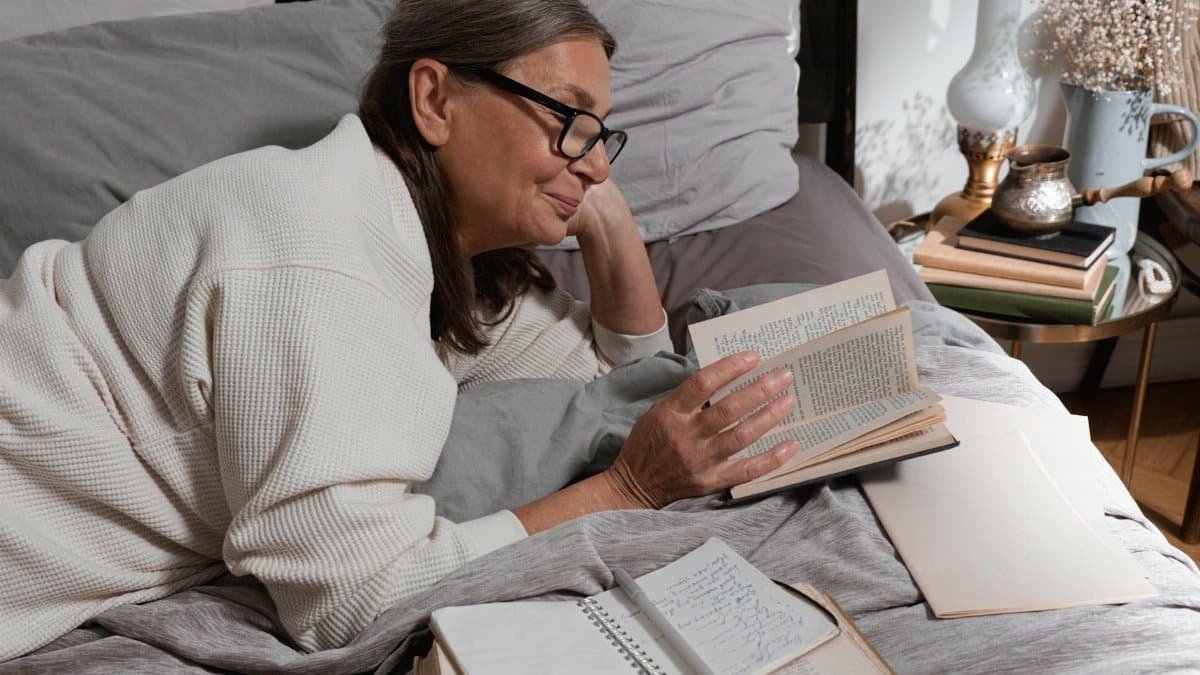 Senior woman reading comfortably in bed with eyeglasses and a cozy blanket, enjoying a serene moment.