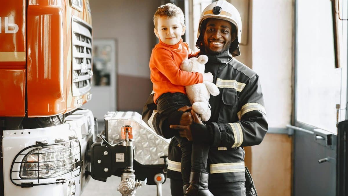 A smiling firefighter holds a boy with a teddy bear at the fire station.