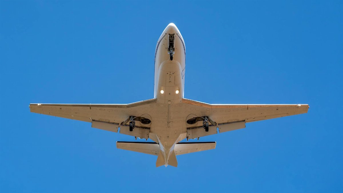 A private jet soaring overhead with landing gear visible against a clear blue sky.