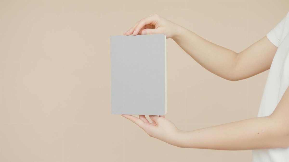 Hands holding a blank book cover for creative design mockup on a neutral background.