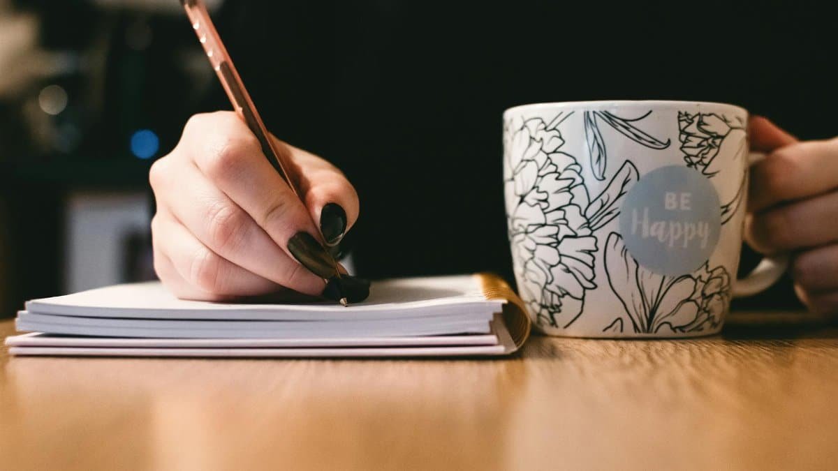 Person writing in a notebook with a floral ceramic mug on a wooden desk.