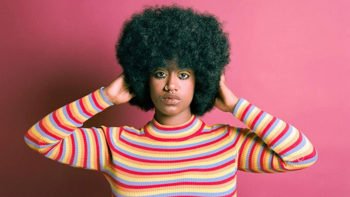Stylish portrait of a woman with afro hair and colorful striped sweater against pink backdrop.