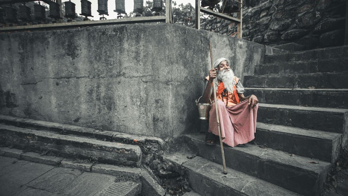 Senior man with white hair and beard sitting on stone stairs, holding a walking stick in a serene outdoor setting.