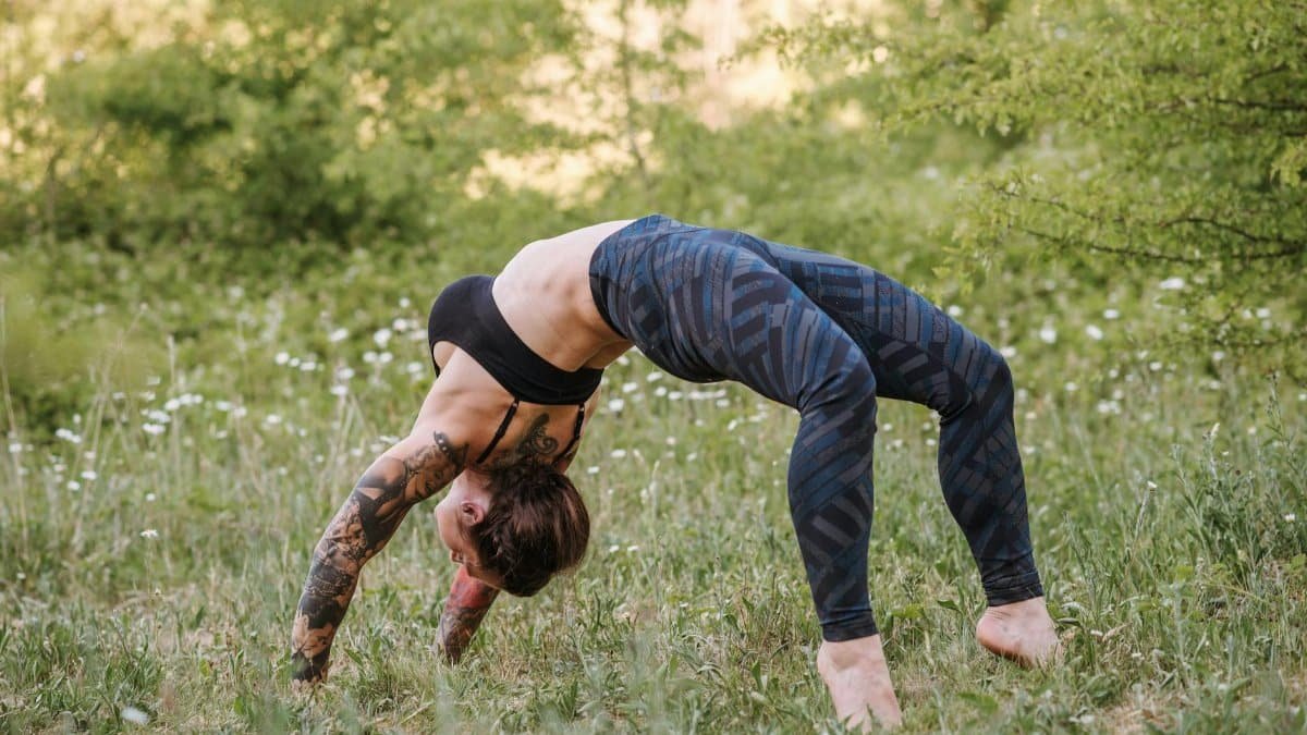 Side view of unrecognizable flexible female in sportswear standing in Urdhva Dhanurasana pose while practicing yoga on meadow