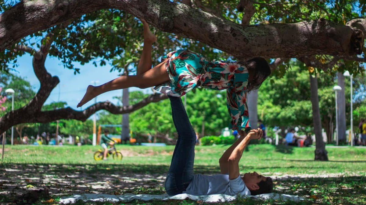 A couple enjoying acro yoga under a tree in a sunny park. Fitness and balance in nature.