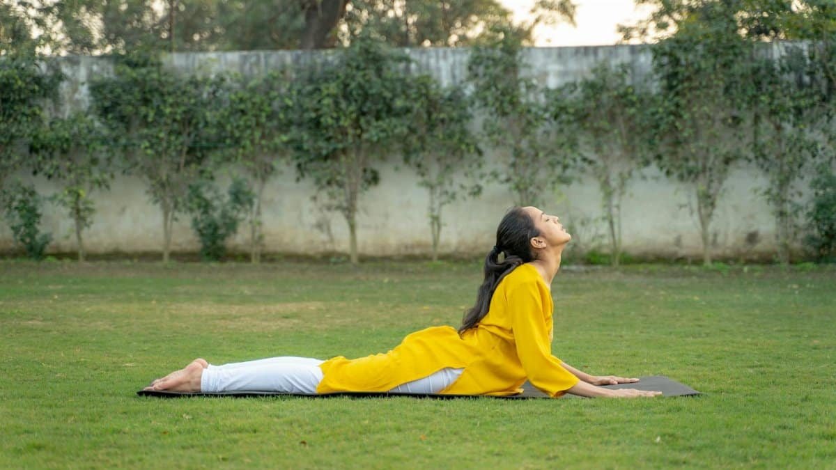 A woman practices yoga outdoors in a serene garden, wearing a yellow shirt and doing the Cobra Pose.