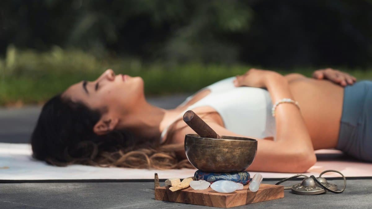 A woman practices relaxation with a singing bowl and crystals in an outdoor setting.