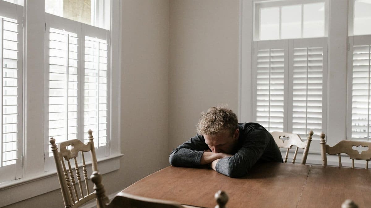 An adult man with emotions of sadness and anxiety sitting alone at a wooden table near windows.