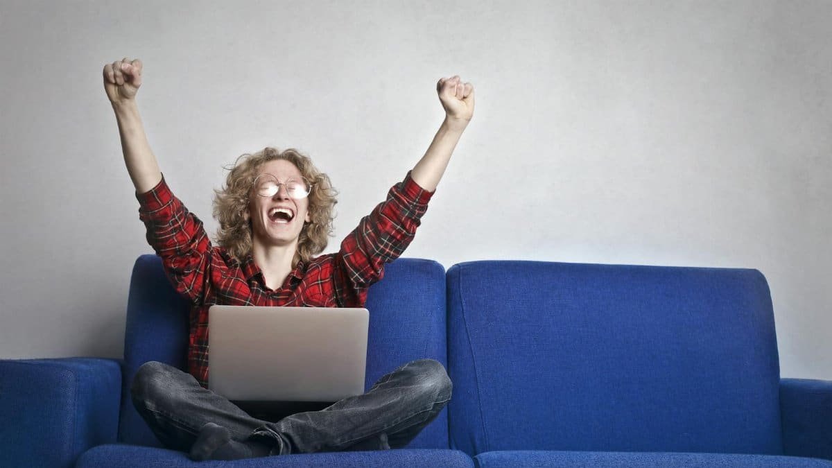 Thrilled individual celebrating success while using a laptop on a blue sofa indoors.