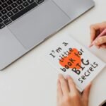 Overhead view of a woman writing in a notebook labeled 'I'm a little book for BIG secrets' beside a laptop.