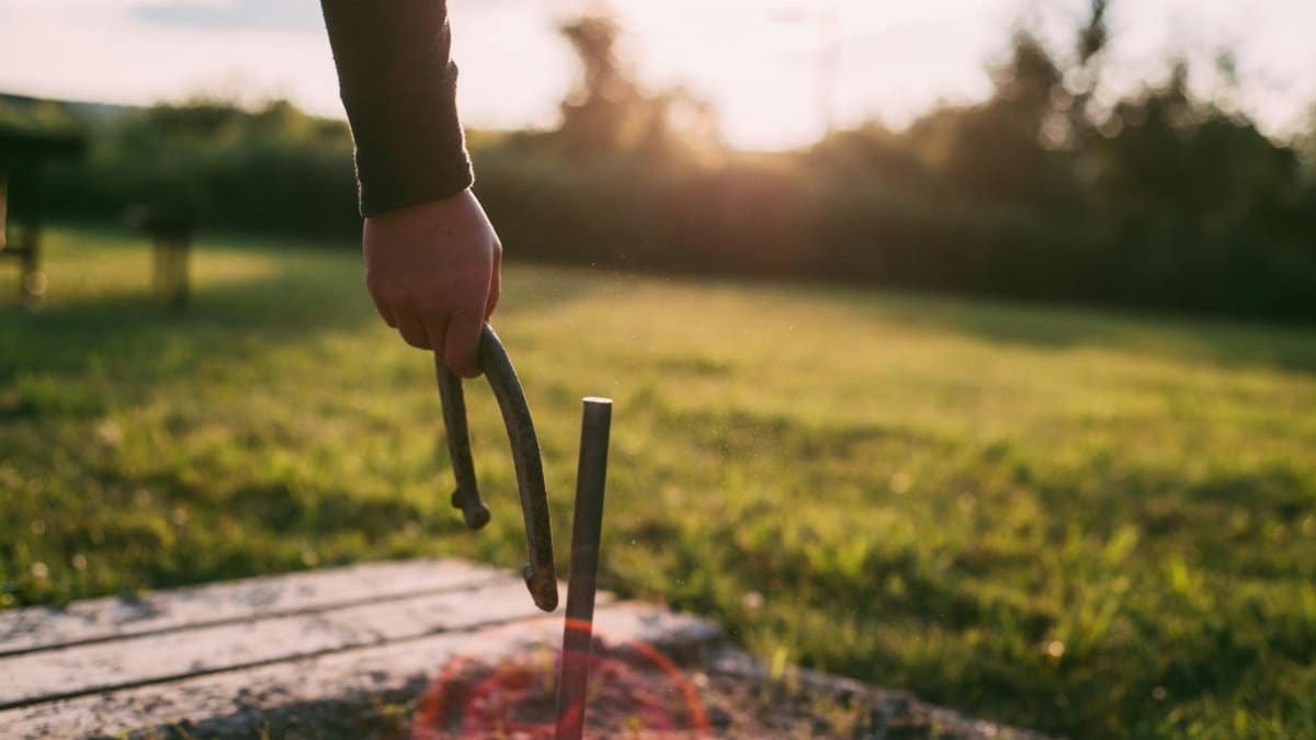 A person's hand throws a horseshoe at a stake on a grassy field during sunset.
