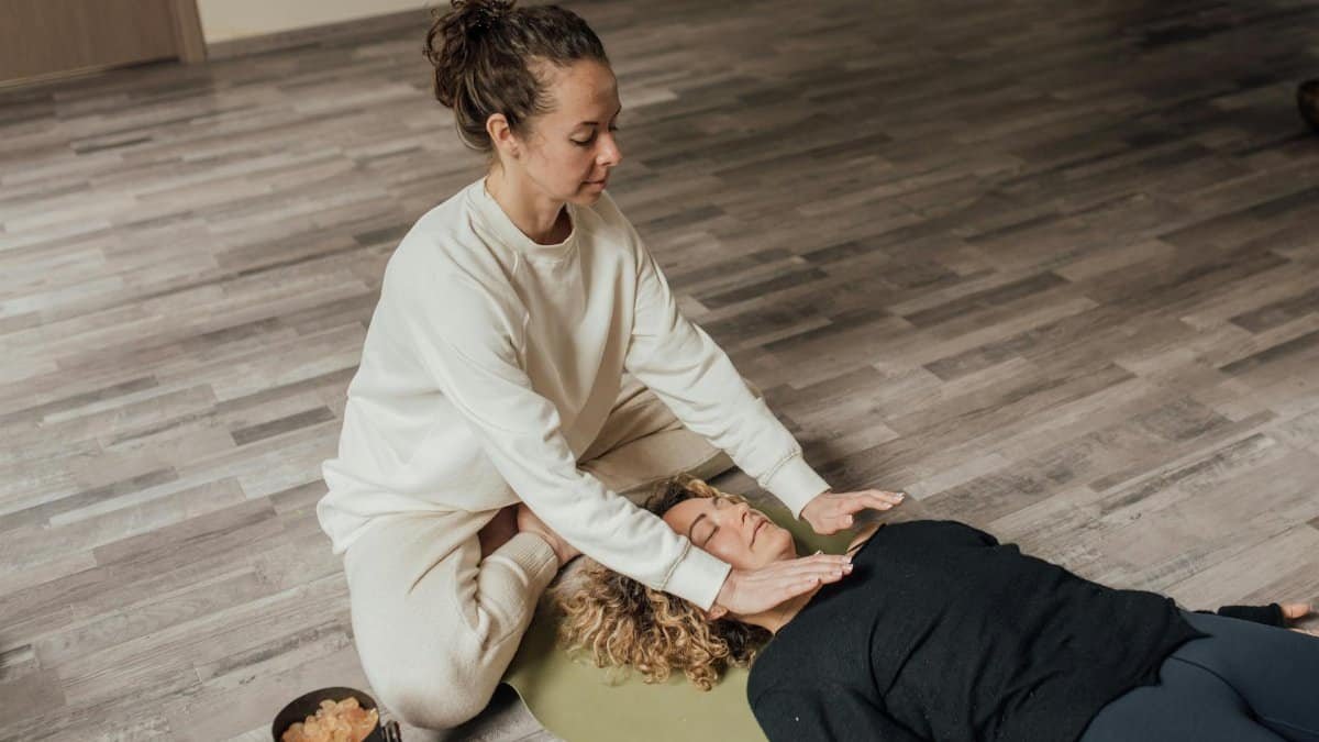 Woman practicing Reiki healing therapy on another woman lying indoors on a yoga mat.