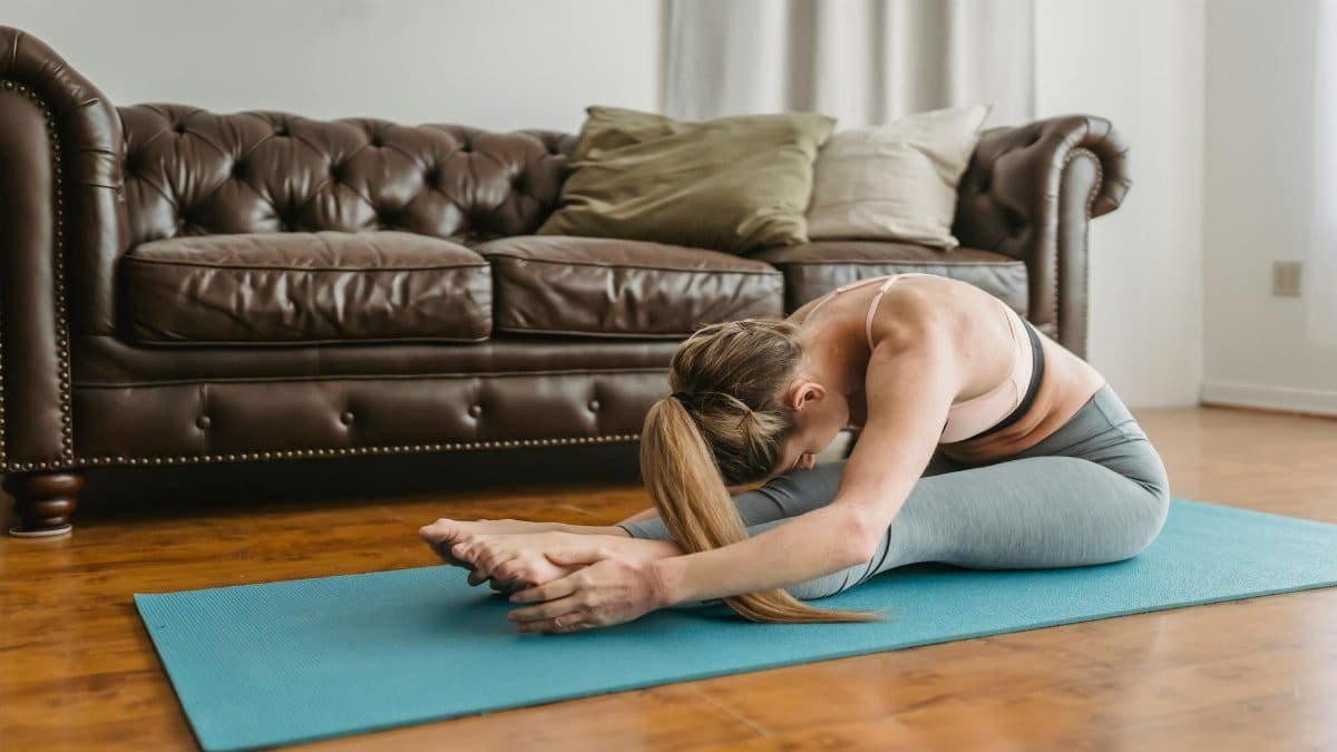 Side view of slim young lady with long blond hair in sportswear recreating in Seated Forward Bend asana after yoga session in living room