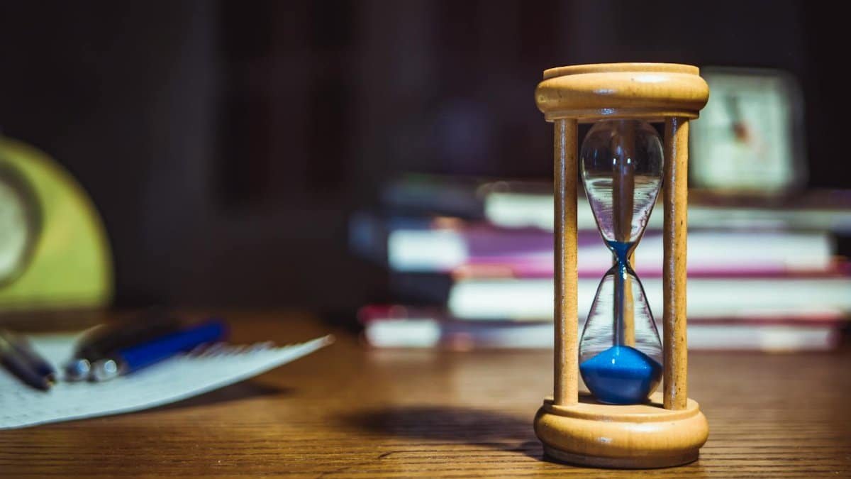 Close-up of a wooden hourglass on a desk, alongside books and pens, suggesting focused study or time management.