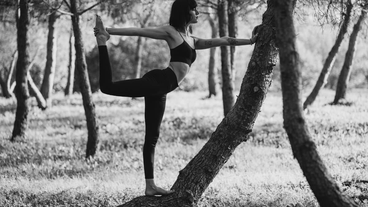 A woman performs the Natarajasana yoga pose surrounded by trees, exemplifying calm and balance.
