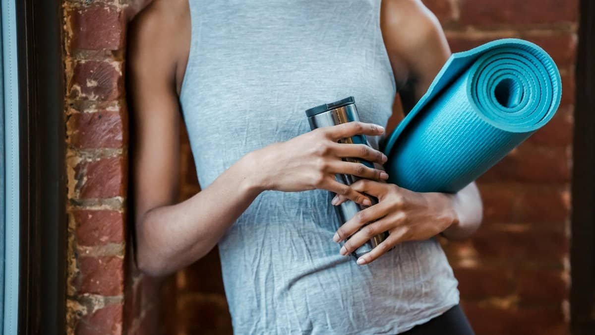 A fit young woman holding a yoga mat and water bottle, standing by a brick wall indoors.
