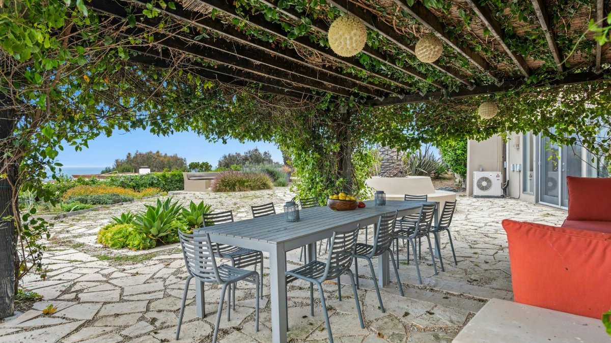 A serene garden patio with a dining setup under a vine-covered pergola, offering an ocean view.