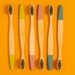 Six bamboo toothbrushes with colorful tips on a vibrant orange surface.