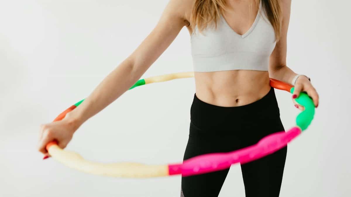 Crop faceless fit lady in black leggings and sports bra practicing fitness exercises with hula hoop against gray background