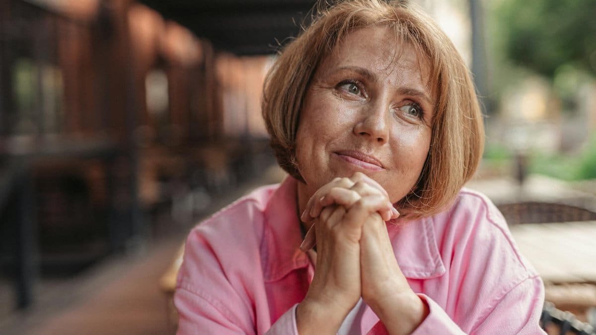 Senior woman with short brunette hair and pink shirt, hands clasped, looking thoughtful outdoors.