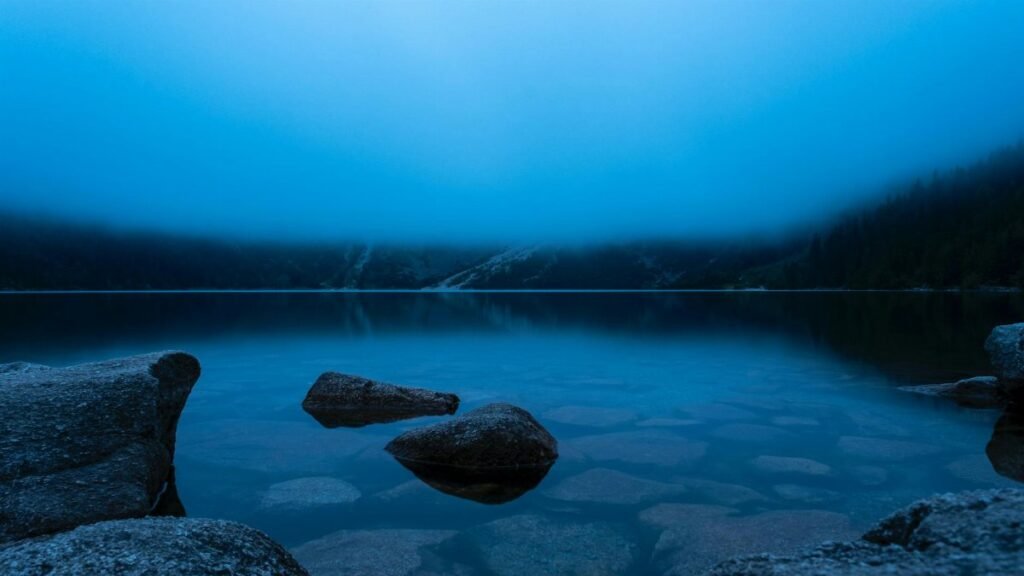 Calm mountain lake shrouded in fog, reflecting tranquil water and rocks.