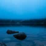 Calm mountain lake shrouded in fog, reflecting tranquil water and rocks.