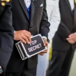 A group of men in formal military and business attire holding a 'Top Secret' briefcase indoors.