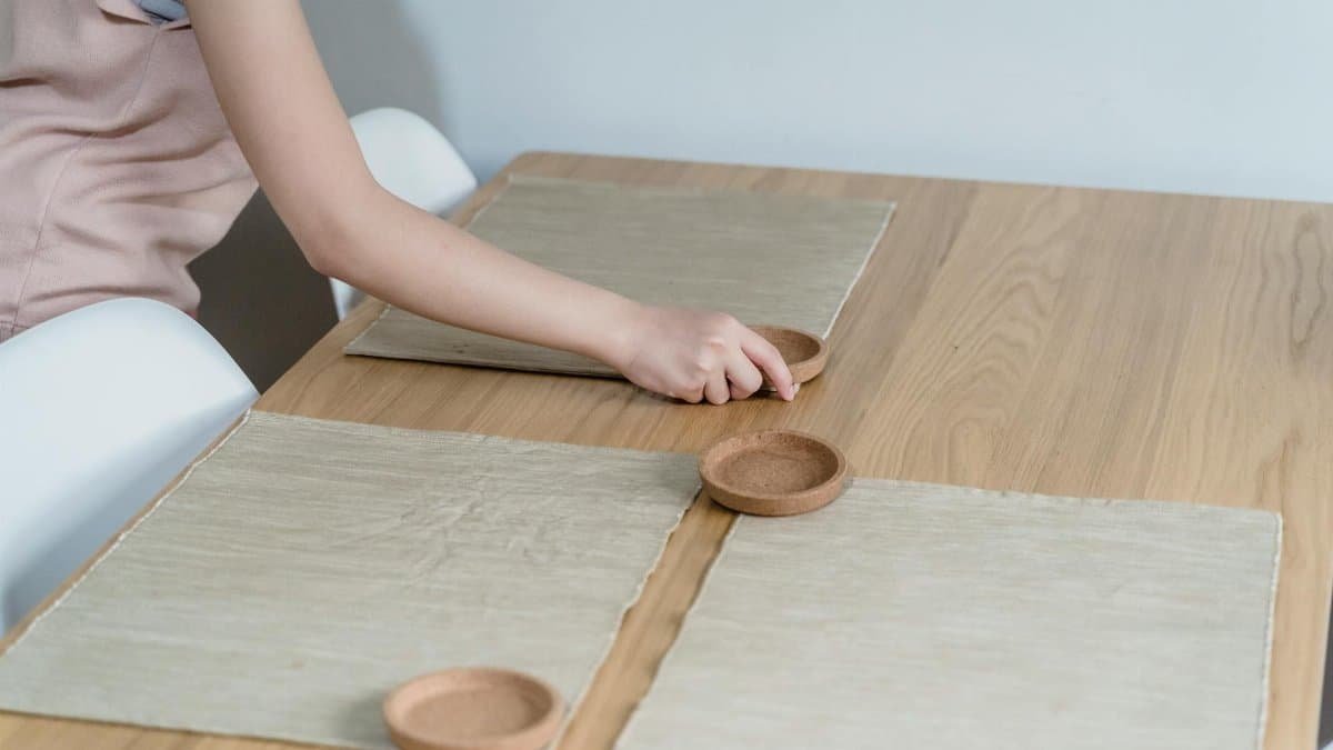 A close-up of a hand arranging a table setting with placemats and coasters on a wooden dining table.