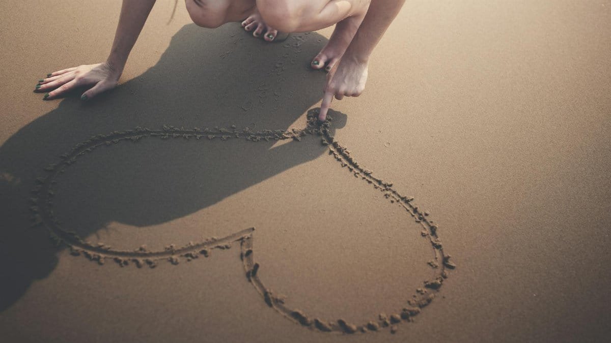 A woman drawing a heart shape in the sand with her finger on a sunny beach day.