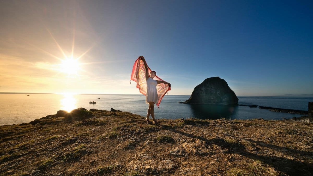 A lone woman with a scarf stands on a rocky cliff, enjoying a stunning ocean sunset view.