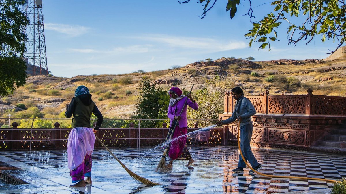 Three individuals cleaning an outdoor tiled courtyard in a scenic Indian landscape under a clear blue sky.