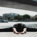 Ground level of anonymous flexible male sitting in Wide Legged Forward Bend pose while practicing yoga near bridge in town