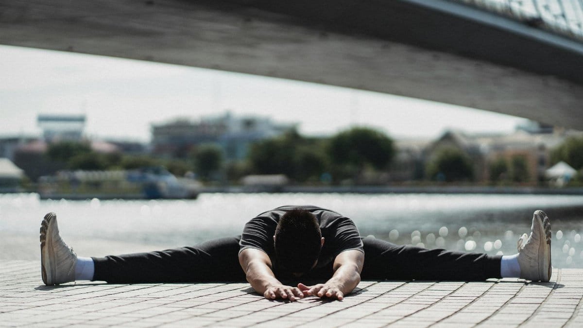 Ground level of anonymous flexible male sitting in Wide Legged Forward Bend pose while practicing yoga near bridge in town