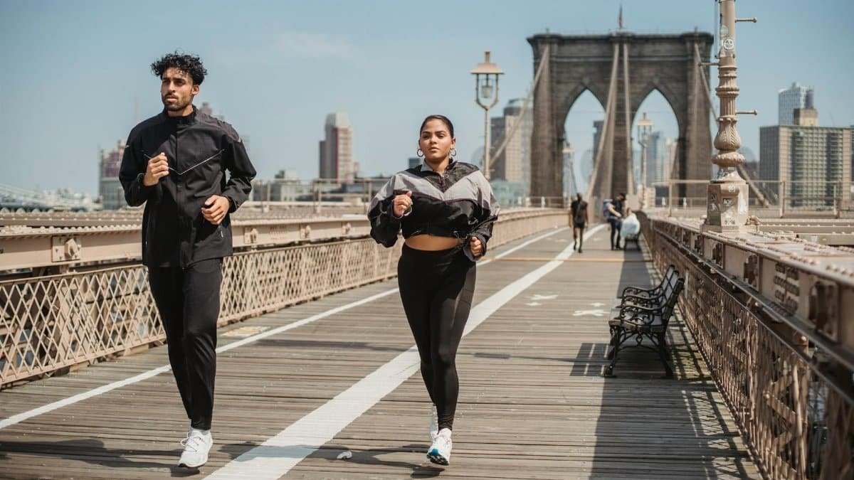 Two adults jogging on Brooklyn Bridge with city skyline in the background, showcasing health and vitality.