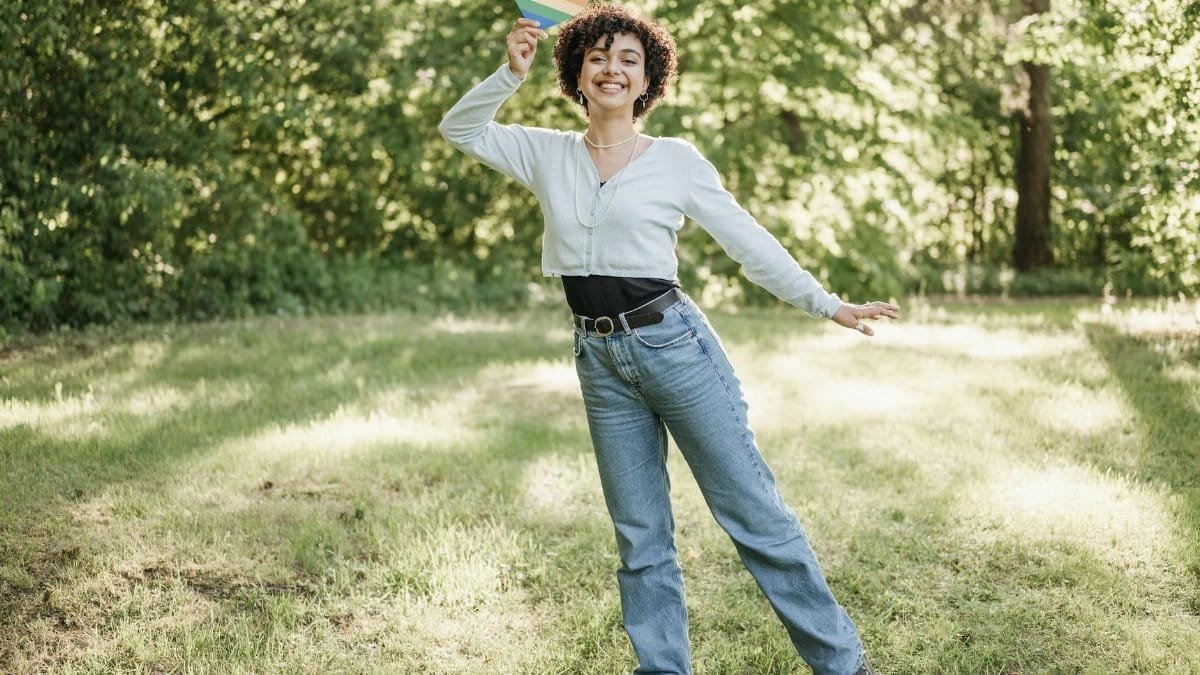 A happy woman with curly hair poses outside with a rainbow heart, embracing Pride.