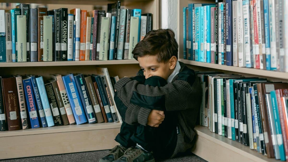 Young boy in library appears sad, sitting alone among bookshelves, suggesting themes of solitude and bullying.
