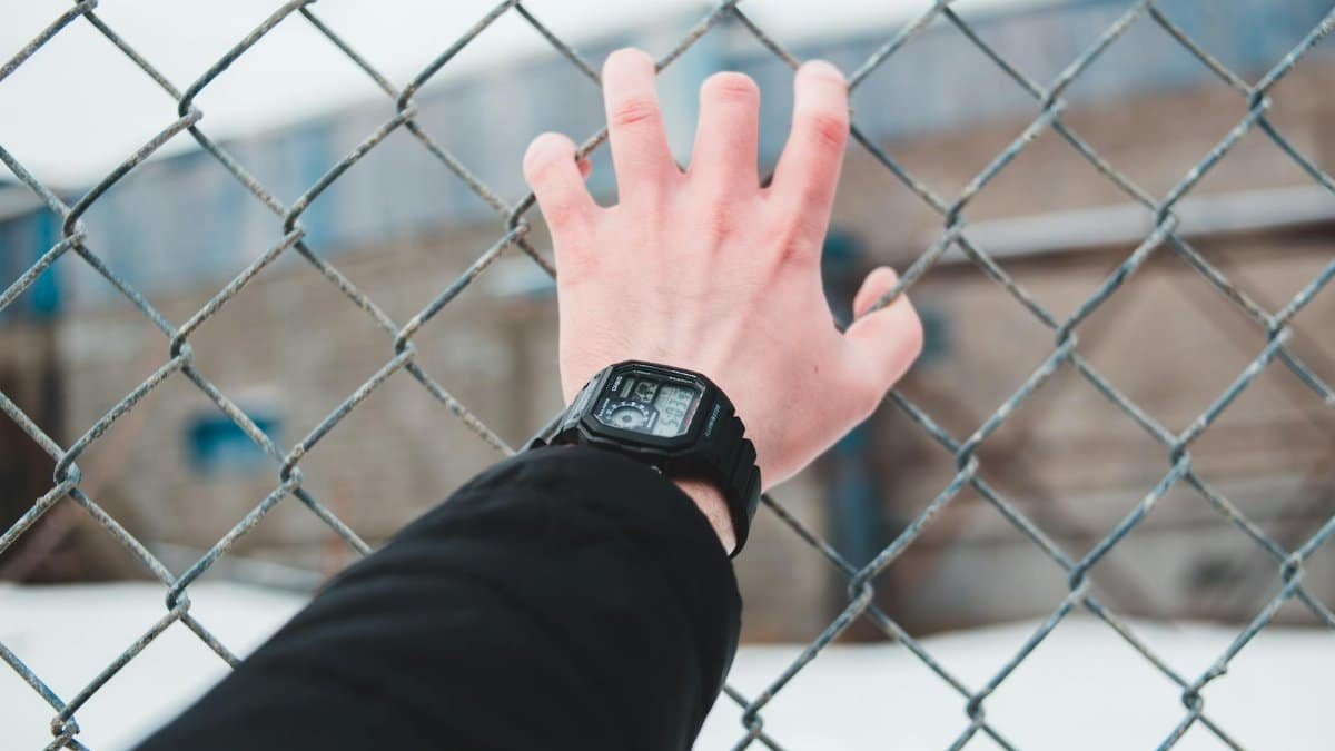 A hand wearing a digital watch reaching through a chain link fence in an outdoor setting.