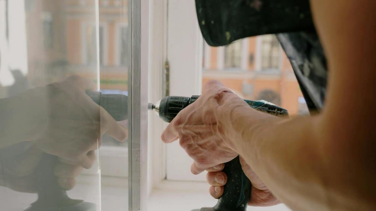 Close-up of a handyman's hands using a drill to fix a window inside an apartment.