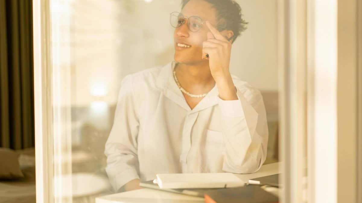Man with glasses sitting indoors, pensive and relaxed. Clearly reflecting through a window with warm lighting.