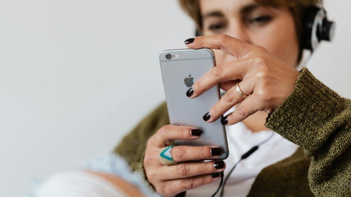 Crop blurred focused female in casual warm clothes with classy ring surfing contemporary cellphone while enjoying favorite music via wired headphones against white plain wall