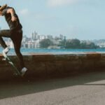 Energetic skateboarder performing tricks near a city waterfront with a clear sky backdrop.