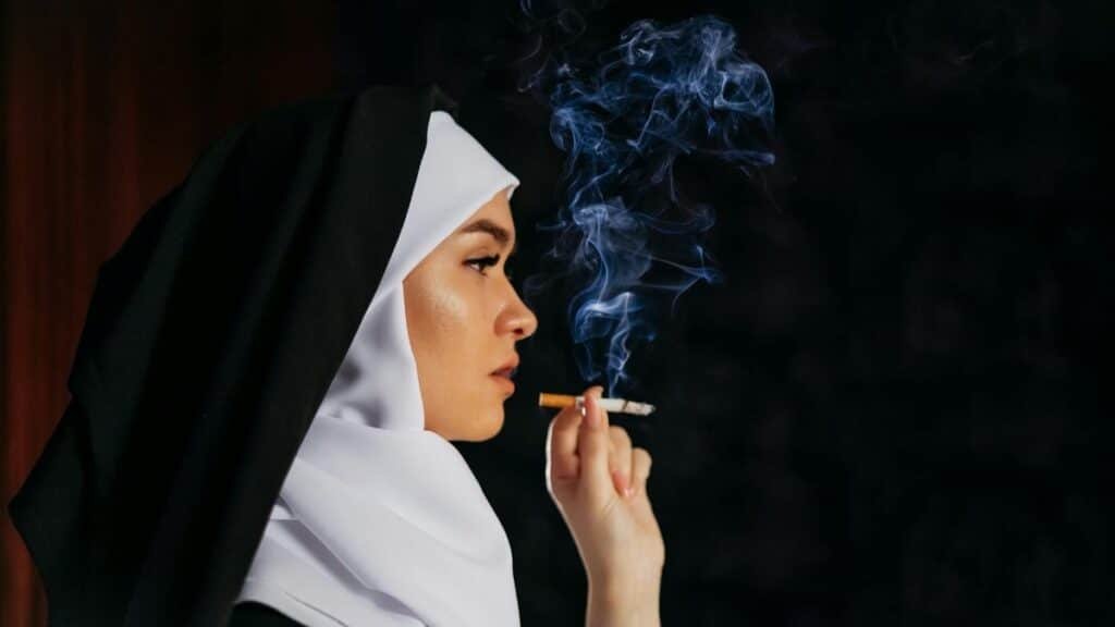Profile of a nun in traditional habit smoking a cigarette, with smoke swirling against a dark backdrop.
