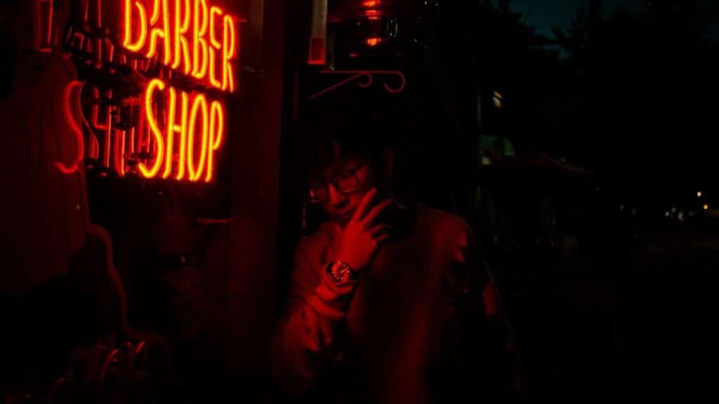 A man stands beside a neon-lit barber shop sign at night, creating a moody atmosphere in Seattle.