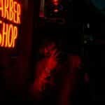 A man stands beside a neon-lit barber shop sign at night, creating a moody atmosphere in Seattle.