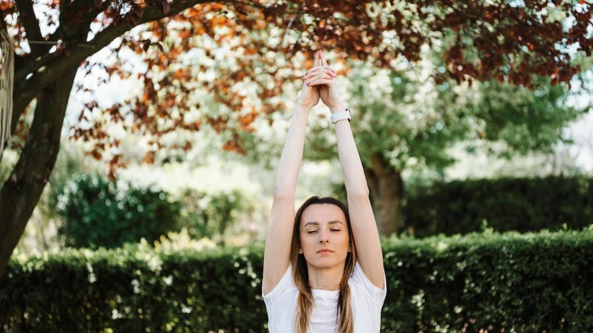 A woman meditating with closed eyes, practicing yoga under a tree in a tranquil outdoor setting.