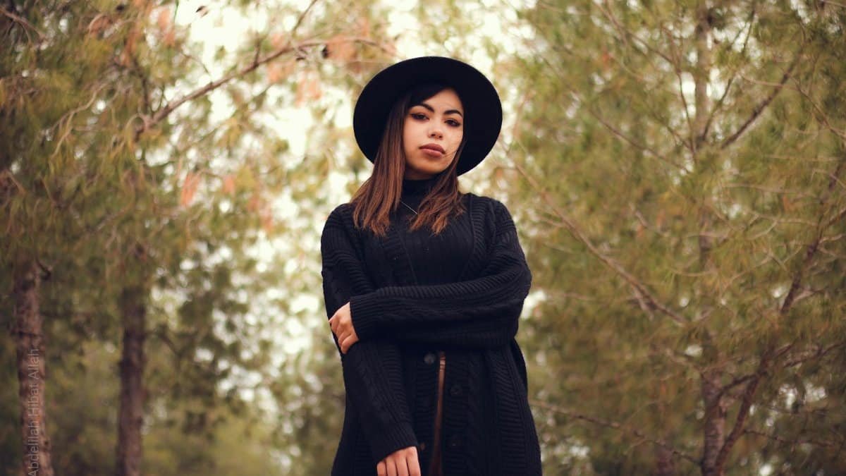 Stylish portrait of a woman in a black hat and sweater, standing in a serene outdoor setting surrounded by trees.