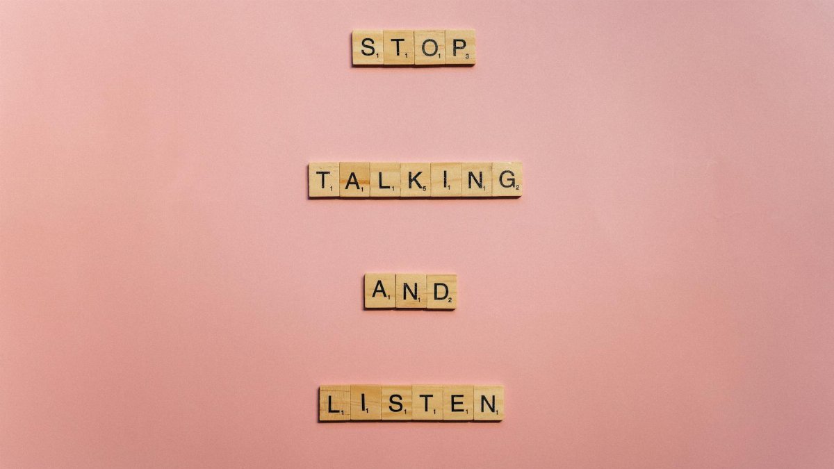 Wooden scrabble tiles form the message 'Stop talking and listen' on a pink surface.